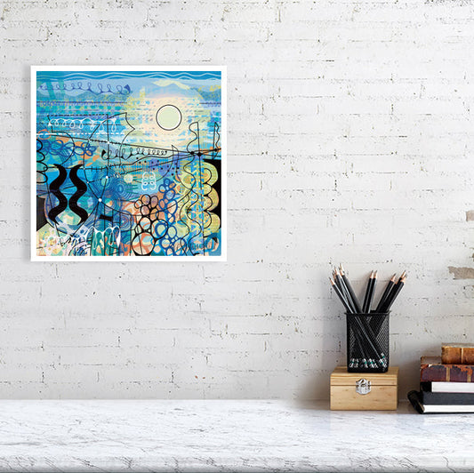 Coastal abstract art print ‘Dazzling’ by Uk artist Ruth Dronsfield shown unframed against a white brick wall above a marble surface with pencils and books. The artwork is in shades of blue and teal wit orange and yellow features and also black and white details. Medium size