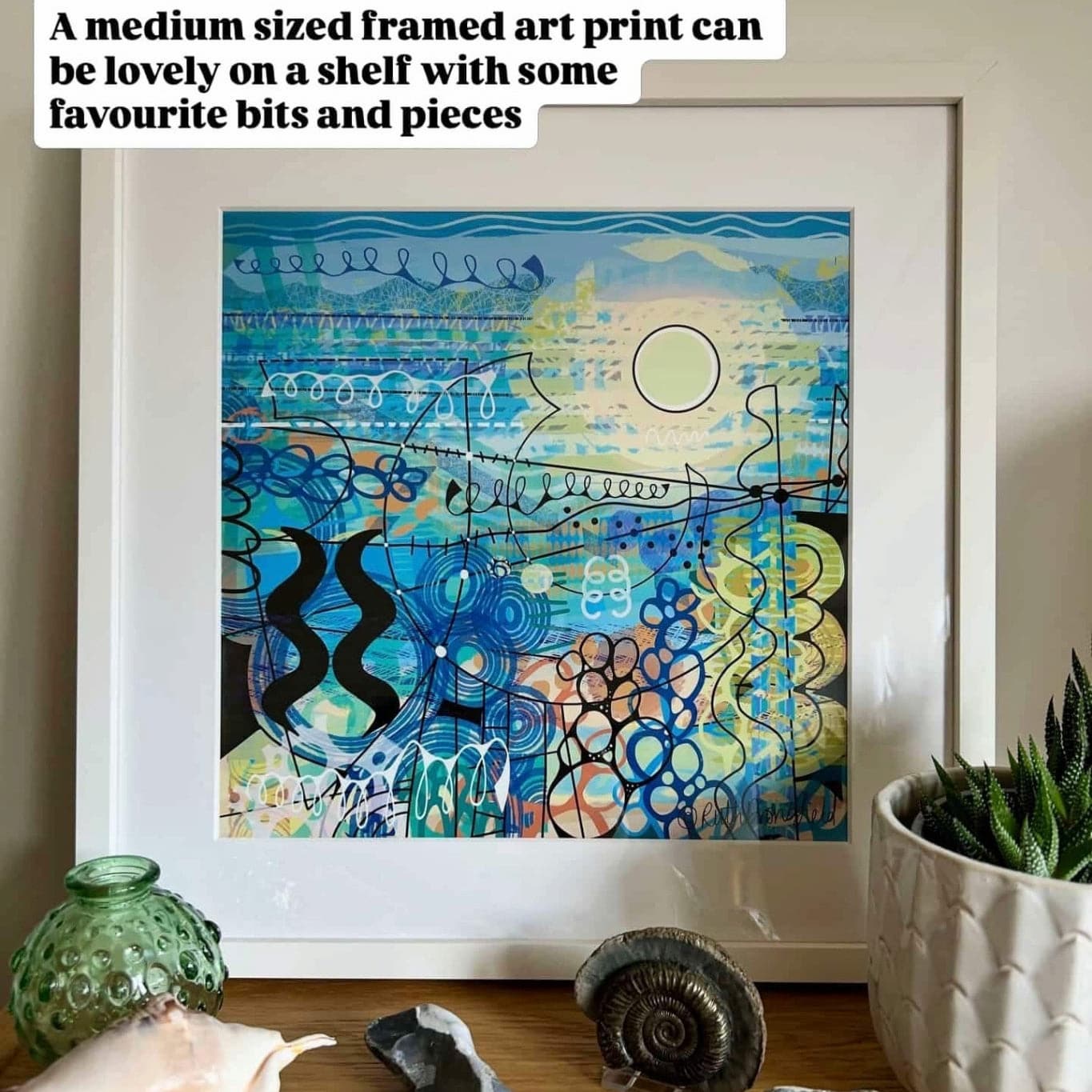 Coastal Abstract Fine Art Print by Uk artist Ruth Dronsfield. Titled DAZZLING. Created in shades of blue with a bright sunshine form and intricate details reminiscent of a pebbly seashore. Shown in a white frame on a shelf
with shells , pebbles, a plant in a white pot and a green glass jar.