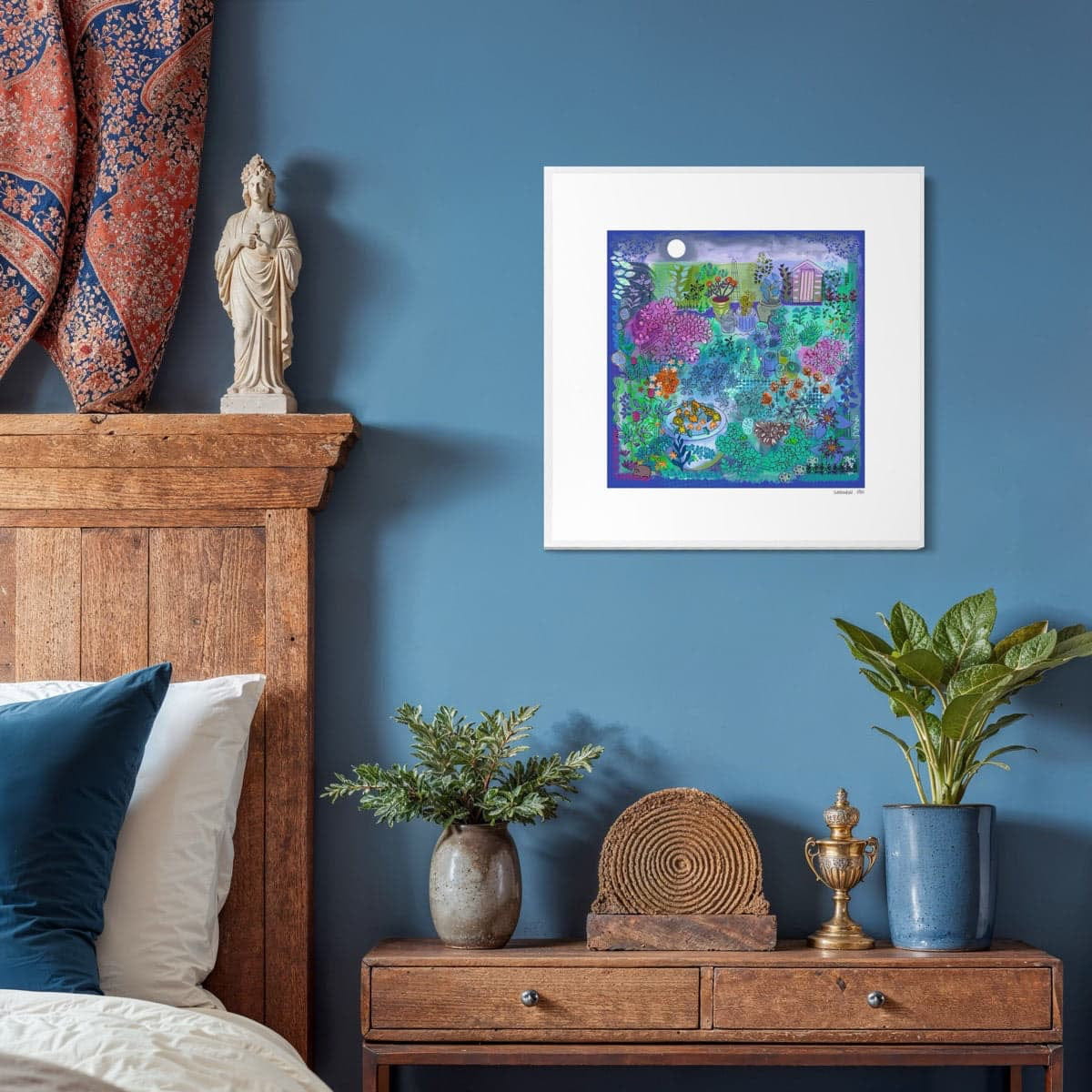 Moonlight Garden-A limited edition fine art giclée print by Ruth Dronsfield UK artist shown here framed in white on a blue wall in a bedroom setting. The artwork has shades of purple blue, green and pink with colourful detail details of plants and wildlife.