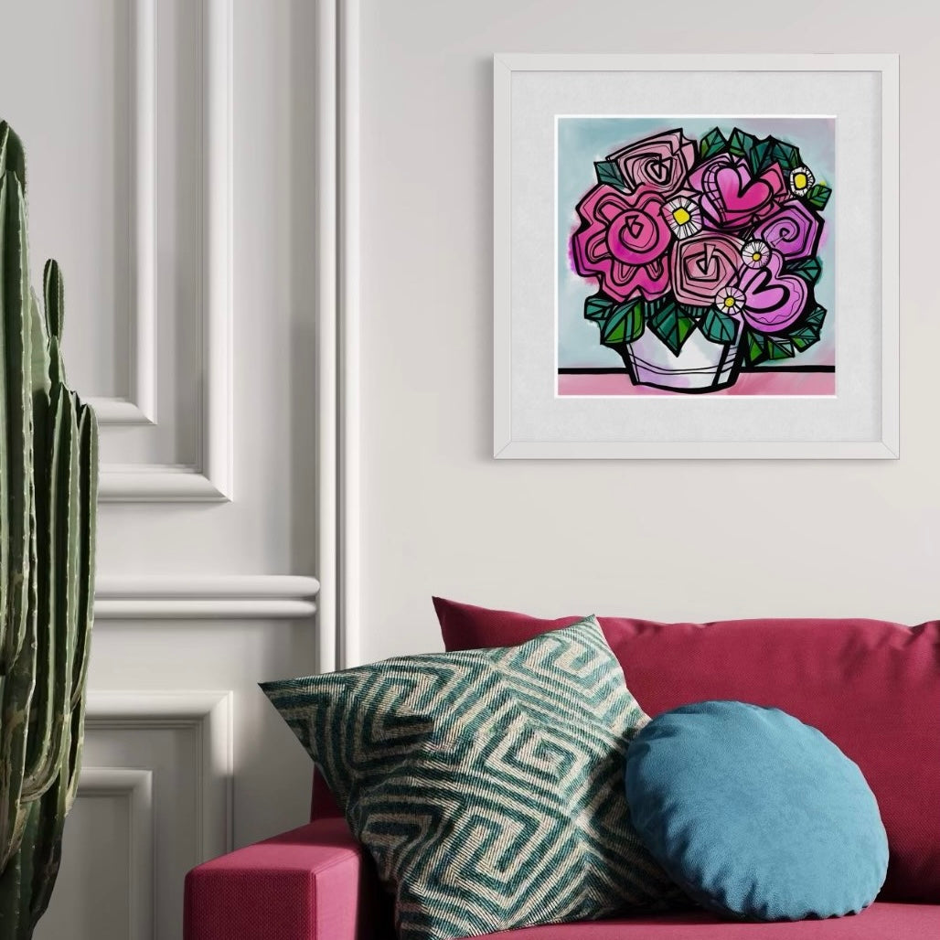 Flower fine art limited edition print by Ruth Dronsfield Art hung framed in white in living room. Artwork of abstract flowers with hearts in shades of pink in a white container on a pink surface with turquoise background. Living room with ruby red sofa with blue and green cushions and a large plant. White wall.