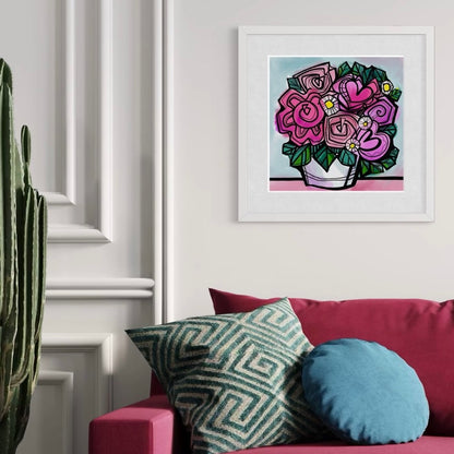 Flower fine art limited edition print by Ruth Dronsfield Art hung framed in white in living room. Artwork of abstract flowers with hearts in shades of pink in a white container on a pink surface with turquoise background. Living room with ruby red sofa with blue and green cushions and a large plant. White wall.