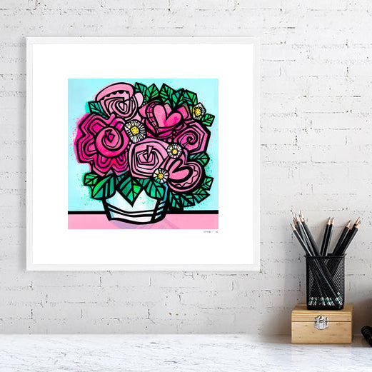 Flower art print from Ruth Dronsfield shown in white frame 40x40cms on a white brick wall above a table with stationary. Flowers in shades of pink and white with yellow details in a white container on a pink surface with turquoise blue background.