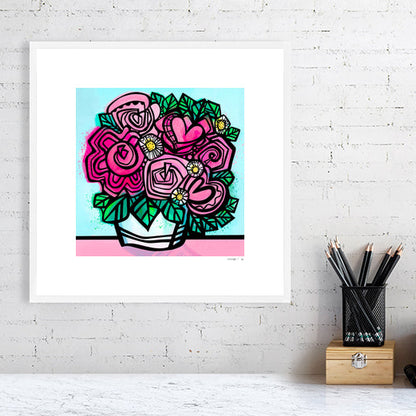 Flower art print from Ruth Dronsfield shown in white frame 40x40cms on a white brick wall above a table with stationary. Flowers in shades of pink and white with yellow details in a white container on a pink surface with turquoise blue background.