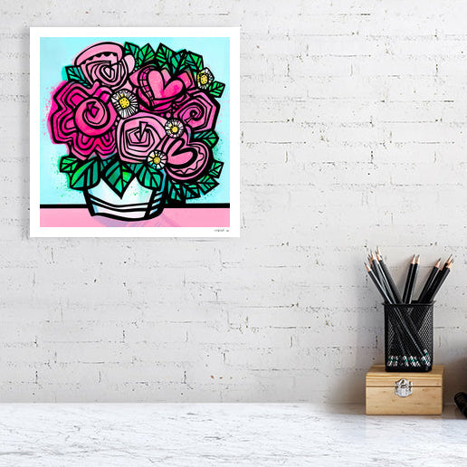 Flower art print from Ruth Dronsfield shown unframed 30x30 cms on a white brick wall above a table with stationary. Flowers in shades of pink and white with yellow details in a white container on a pink surface with turquoise blue background.