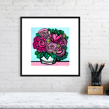 Flower art print from Ruth Dronsfield shown in a black frame 40x40cms on a white brick wall above a table with stationary. Flowers in shades of pink and white with yellow details in a white container on a pink surface with turquoise blue background.