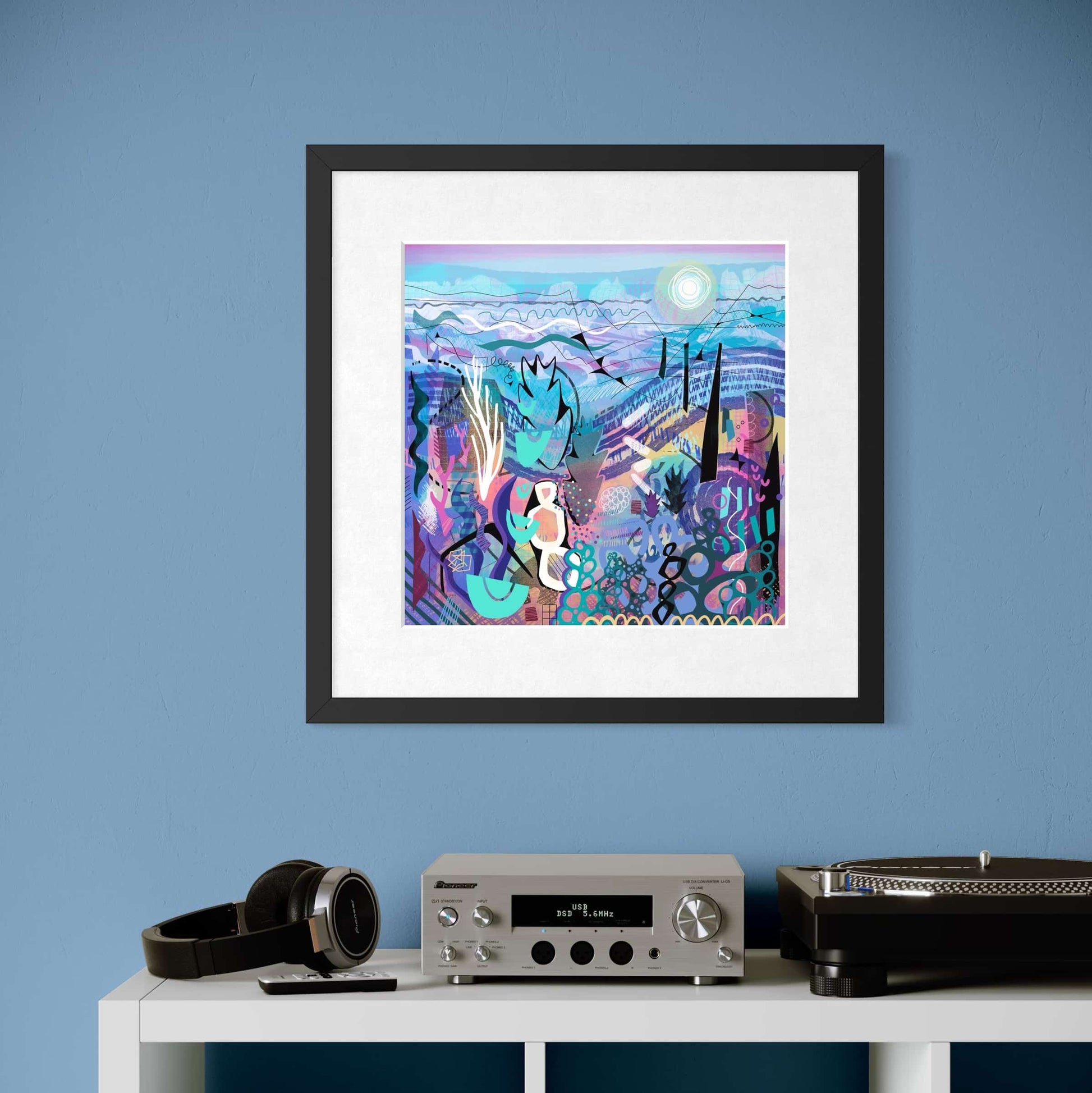 Framed abstract artwork print by UK artist Ruth Dronsfield on a blue wall above a white shelf with electronic equipment and books.