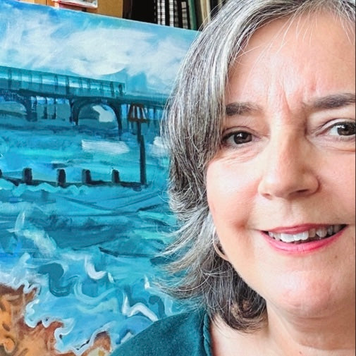 UK artist Ruth Dronsfield  standing next to a painting of a coastal scene with a pier.