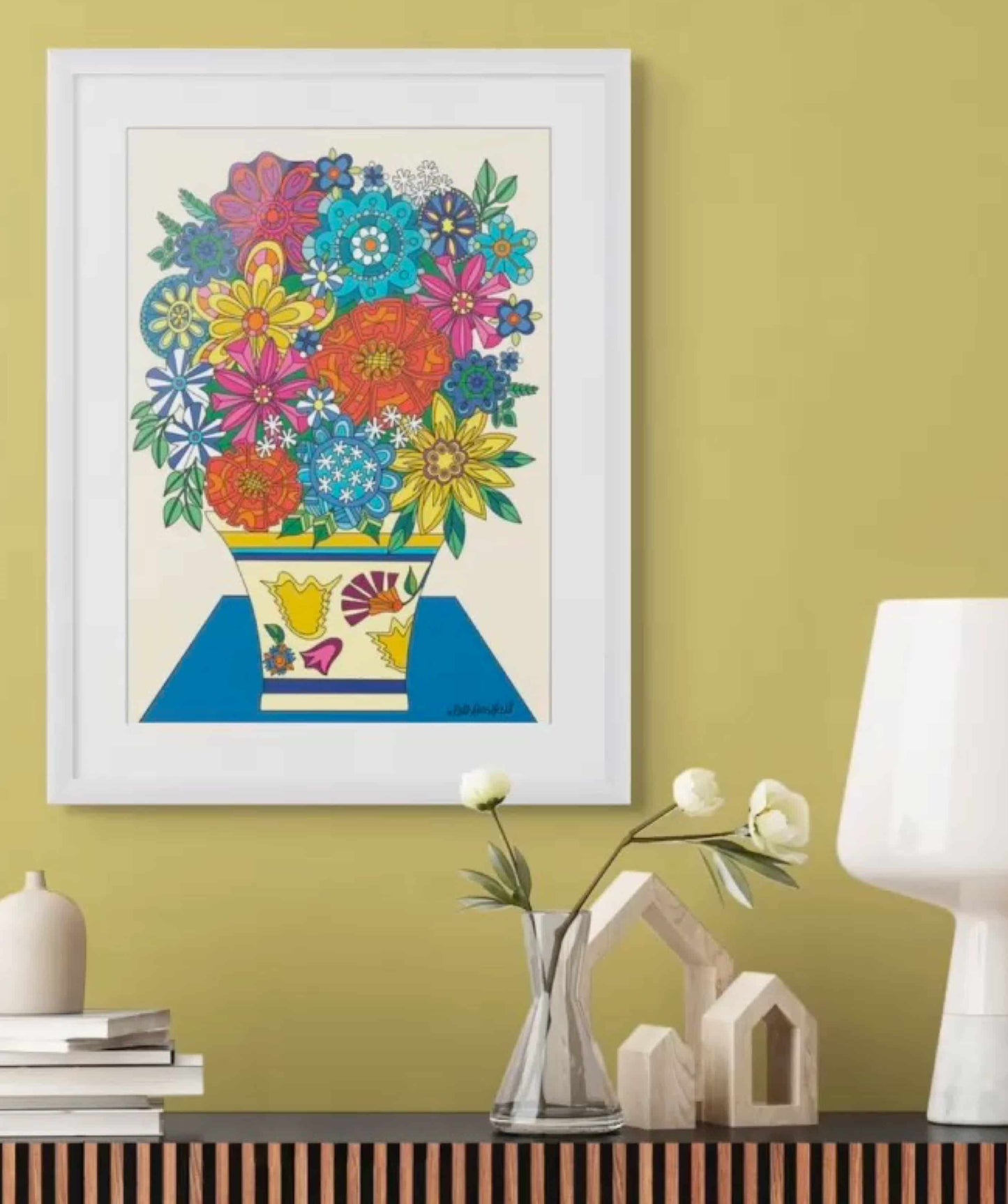 Colorful floral print framed in white on a yellow wall with decorative items below.